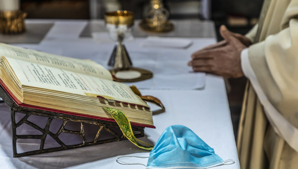 Ein Priester faltet die Hände und feiert den Gottesdienst am Altar, auf dem das Messbuch aufgeschlagen liegt, daneben eine einfache Atemschutzmaske, dahinter liturgische Geräte